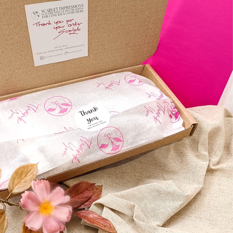 May include: An open cardboard box with a pink lid, revealing white tissue paper with a pink butterfly design and the text "Thank You". The tissue paper is inside a brown box with a white label that says "Scarlet Impressions".