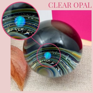 Pode incluir: Uma esfera de vidro transparente com um opala azul embutido no centro. A esfera &eacute; cercada por camadas de vidro preto, dourado e branco, criando um padr&atilde;o de redemoinho. A esfera est&aacute; sentada em uma superf&iacute;cie de tecido marrom claro.
