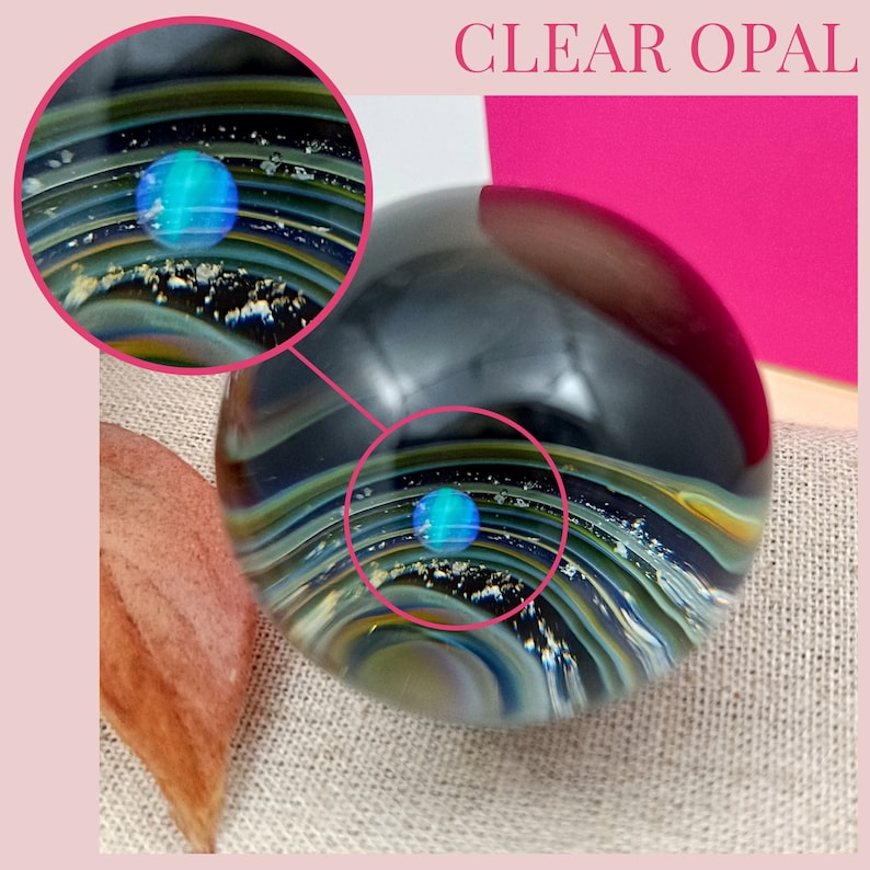 Pode incluir: Uma esfera de vidro transparente com um opala azul embutido no centro. A esfera &eacute; cercada por camadas de vidro preto, dourado e branco, criando um padr&atilde;o de redemoinho. A esfera est&aacute; sentada em uma superf&iacute;cie de tecido marrom claro.