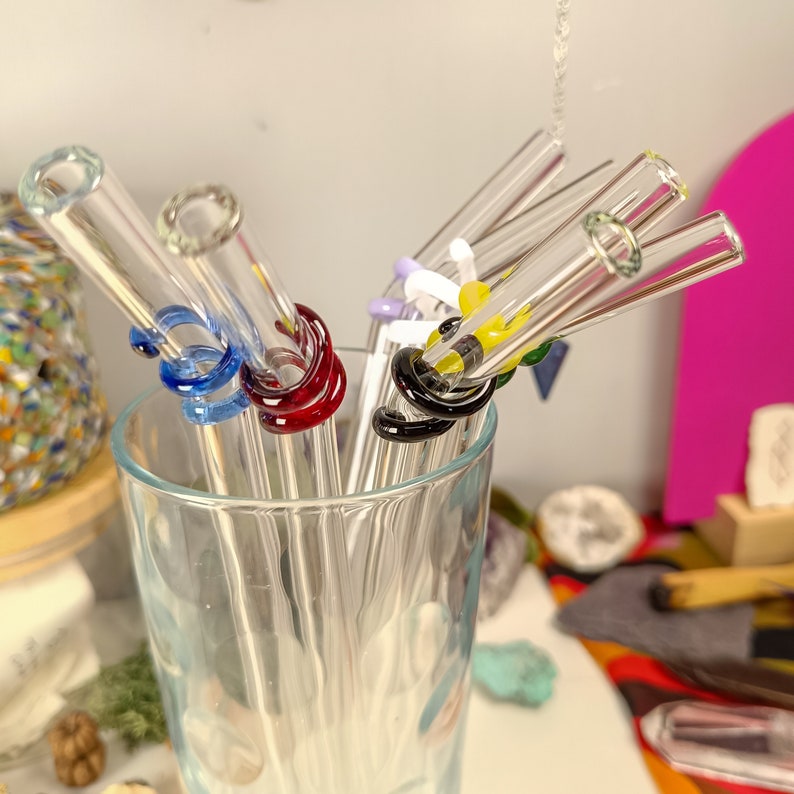 May include: A set of colorful glass drinking straws with decorative glass rings. The straws are arranged in a glass, and the rings are in various colors, including blue, red, yellow, and black.