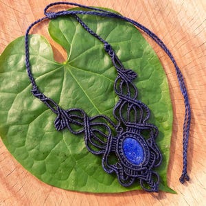 Könnte beinhalten: Dunkelblaue Makramee-Halskette mit einem ovalen, blauen Stein. Die Kette liegt auf einem großen grünen Blatt, vor einem Holzuntergrund. Die aufwendige Knotenarbeit der Halskette ist deutlich sichtbar.