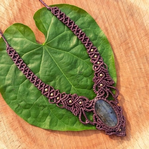 May include: A handcrafted macramé necklace in purple and gold tones, with an oval, iridescent gemstone. The necklace is presented on a large green leaf, set against a wooden background.