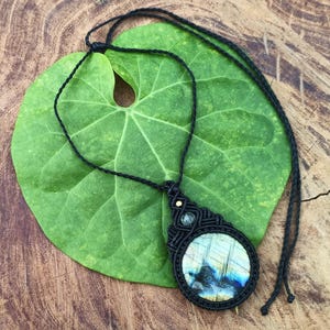 以下が含まれることがあります： 青と黄色の色合いの、丸い虹色の石のペンダントが付いた手作りのマクラメネックレス。ペンダントは黒いマクラメ織りで囲まれ、黒いコードから吊り下げられています。ネックレスは、木の背景に対して緑の葉の上に置かれています。