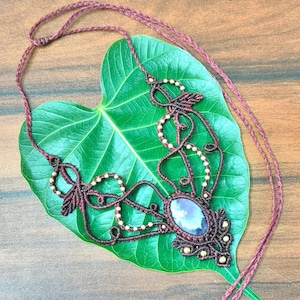 Op de afbeelding: Handgemaakte macramé ketting in bordeauxrood met een ovale steen en kleine goudkleurige kralen. De ketting ligt op een groot groen blad op een houten achtergrond.