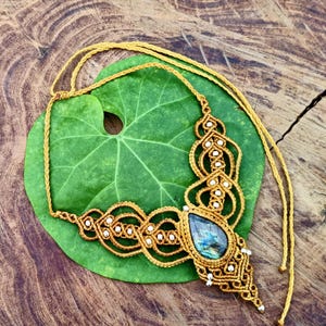 Può includere: Una collana macramè fatta a mano con un ciondolo a forma di goccia di pietra preziosa iridescente. La collana presenta un'intricata annodatura in un colore marrone dorato, accentuata da piccole perle bianche. La collana è esposta su una grande foglia verde.