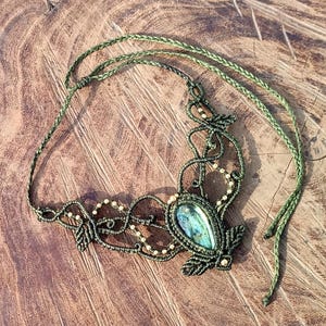 May include: A handcrafted macrame necklace with a central teardrop-shaped gemstone. The necklace features intricate knotting in dark green cord, accented with small, light-colored beads. The gemstone has a blue-green iridescence.
