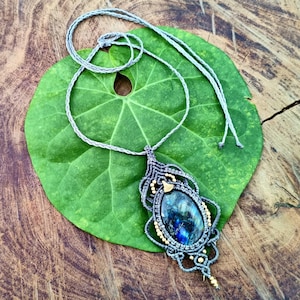Può includere: Una collana macramè fatta a mano con un grande pendente ovale di pietra preziosa blu e verde. Il pendente è intrecciato in modo intricato con fili grigi e dorati. La collana è esposta su una foglia verde su uno sfondo di legno.