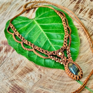 Op de afbeelding: Handgemaakte macramé ketting met een bruin koord, met een ovale edelsteen hanger. De ketting ligt op een groot groen blad, tegen een houten achtergrond. De ingewikkelde knoop- en kralenwerk creëert een bohemien esthetiek.