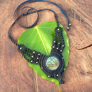 以下が含まれることがあります： 中央に丸い虹色の石をあしらった、手作りのマクラメネックレス。 ネックレスは黒色で、金色と濃い青色のコードがアクセントになっています。 大きな緑色の葉の上に置かれ、木製の背景になっています。