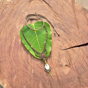 Può includere: Una collana macramè fatta a mano con un cordoncino marrone e un ciondolo a conchiglia. La collana è esposta su una grande foglia verde, su uno sfondo di legno rustico. La collana presenta piccole perline color oro.