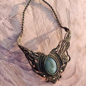 Peut inclure: Collier macramé fait main avec un pendentif ovale bleu-vert. Le cordon vert foncé est noué de manière complexe, formant un motif décoratif. De petites perles métalliques ornent le design. Un bijou artisanal de style bohème.