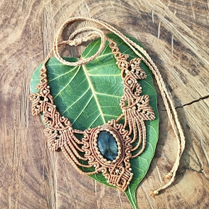 May include: A handcrafted macrame necklace with a central oval-shaped gemstone. The necklace features intricate knotting in a light brown color, with a matching cord. The necklace is displayed on a large green leaf, set against a wooden background.