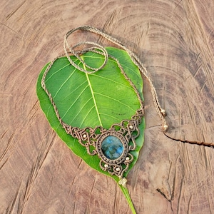 Może przedstawiać: Ręcznie robiony naszyjnik z makramy z centralnym niebiesko-zielonym kamieniem szlachetnym. Naszyjnik ma skomplikowane węzły w brązowym sznurku, z małymi białymi koralikami. Naszyjnik jest prezentowany na dużym zielonym liściu na drewnianym tle.