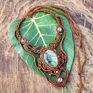 May include: A handcrafted macrame necklace featuring a central oval-shaped stone with a blue-green hue, surrounded by intricate brown macrame knots. Clear beads and gold accents are incorporated into the design. The necklace rests on a green leaf.