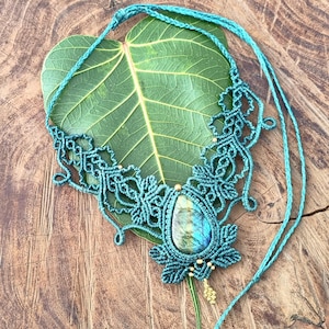 Könnte beinhalten: Eine türkisfarbene Makramee-Halskette mit einem tropfenförmigen Edelsteinanhänger. Das aufwendige Makramee-Design weist florale Elemente und kleine goldene Perlen auf. Die Halskette wird auf einem grünen Blatt vor einem Holzgrund präsentiert.