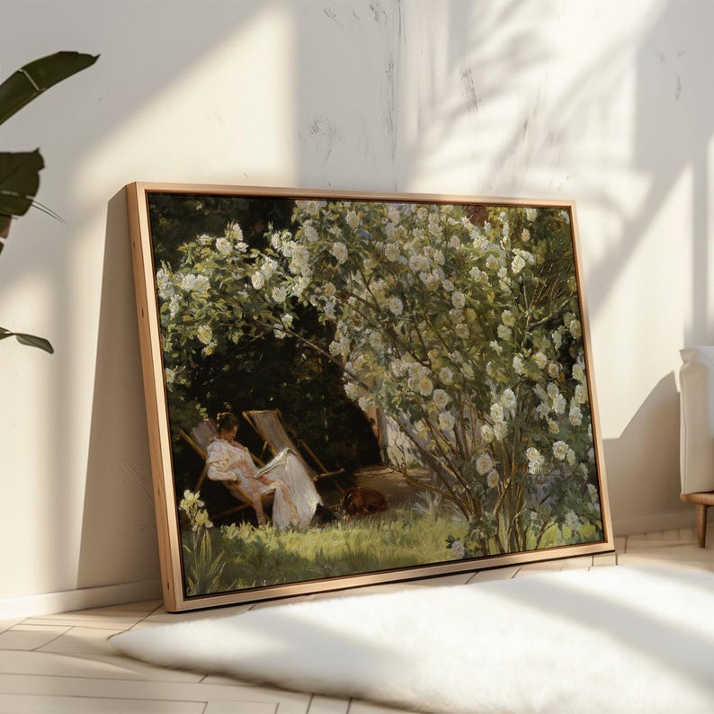 May include: A framed art print of a woman reading in a garden. The artwork depicts a woman in a white dress seated in a wooden chair under a large flowering bush. The painting has a natural wood frame and is displayed indoors.