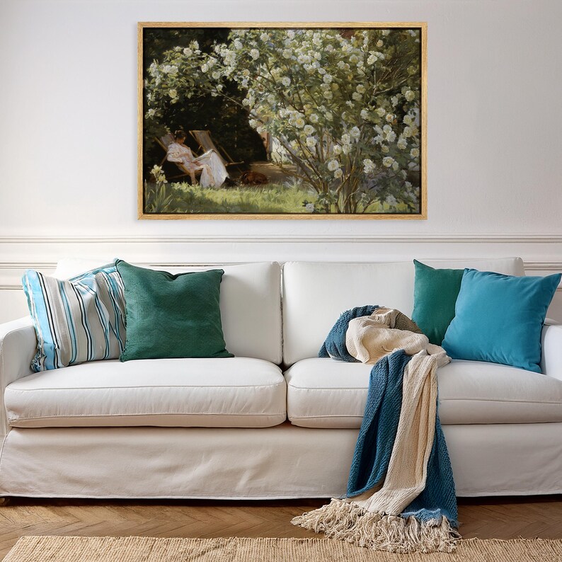 May include: A living room scene featuring a white sofa adorned with colorful throw pillows in shades of blue and green. A framed painting with a gold frame hangs above the sofa. A blue and cream throw blanket with fringe is draped over the sofa.