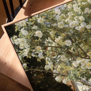 May include: A framed painting featuring a close-up of a white rose bush in full bloom. The artwork has a natural wood frame and is set against a wooden surface. The painting's style is reminiscent of impressionism, with visible brushstrokes and a focus on light and color.