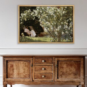 May include: A framed painting depicts a woman in a white dress relaxing in a chair under a flowering bush. The artwork, in a gold frame, is set against a backdrop of lush greenery and a sunny outdoor scene. A dog rests near the woman.