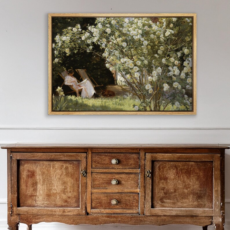 May include: A framed painting depicts a woman in a white dress relaxing in a chair under a flowering bush. The artwork, in a gold frame, is set against a backdrop of lush greenery and a sunny outdoor scene. A dog rests near the woman.