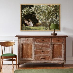 May include: A framed painting depicts a woman reading in a garden, with white flowers and a dog. The painting hangs above a distressed wooden cabinet with drawers and doors. A green lamp sits on the cabinet, and a wooden chair with a green cushion is in the foreground.