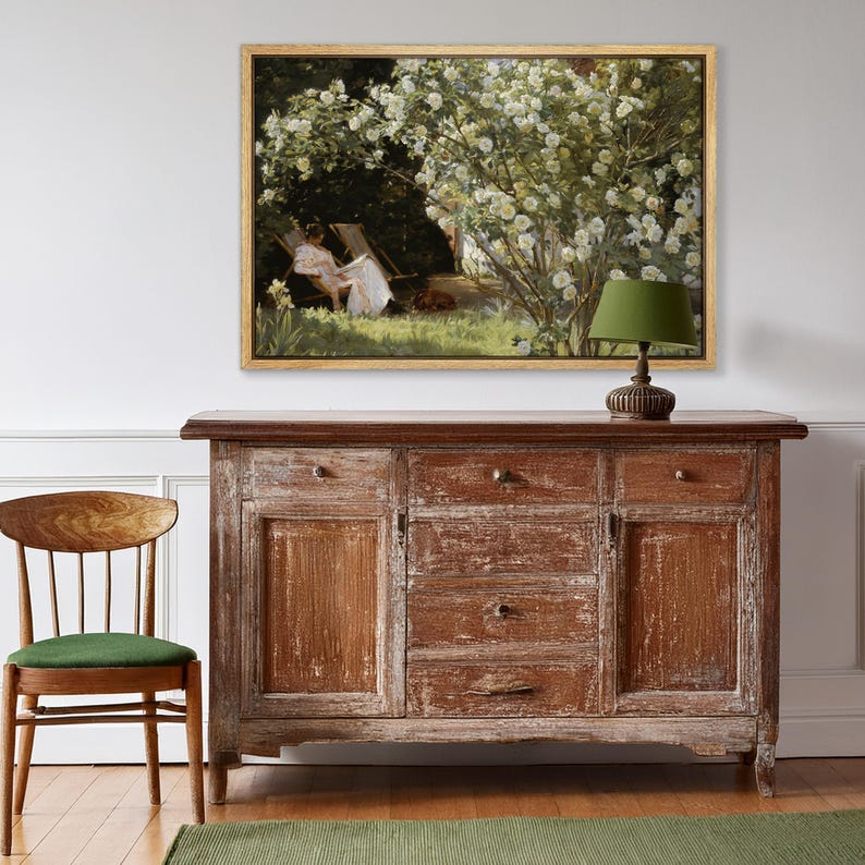 May include: A framed painting depicts a woman reading in a garden, with white flowers and a dog. The painting hangs above a distressed wooden cabinet with drawers and doors. A green lamp sits on the cabinet, and a wooden chair with a green cushion is in the foreground.