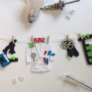 May include: A felt garland with science-themed decorations, including a beaker, a lab coat, a microscope, a molecule, and a test tube. The garland is strung on a white string with clothespins.