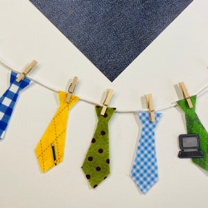Peut inclure: Une guirlande de huit cravates en feutre, chacune avec un motif différent, suspendues à des pinces à linge sur une ficelle blanche. Les cravates sont vertes avec des étoiles blanches, à carreaux bleus et blancs, jaunes avec un motif diagonal, vertes avec des pois marrons, à carreaux bleus et blancs, vertes avec un graphique d'ordinateur portable, rouges et blanches à rayures, et vertes avec une étoile blanche.