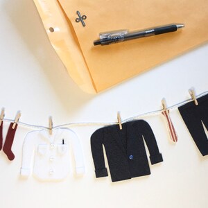 Peut inclure: Une chaîne de vêtements en feutre, comprenant une chemise, un pantalon, une cravate et une paire de chaussures, sont accrochés à une corde à linge avec des pinces à linge. Les articles sont tous blancs, à l'exception du pantalon et de la cravate, qui sont respectivement noirs et rouges. La corde est suspendue sur un fond blanc. Le texte "#1 DAD" est visible sur l'un des articles en feutre.