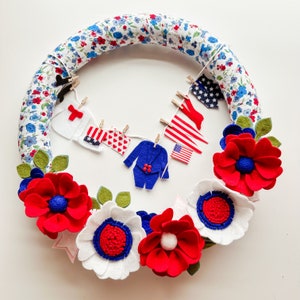 May include: A patriotic wreath with red, white, and blue felt flowers and a garland of felt clothing items. The wreath is made of a white fabric with a floral pattern.