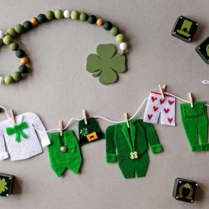 Puede incluir: Una guirnalda de fieltro verde con una variedad de artículos temáticos del Día de San Patricio, que incluyen un trébol, un sombrero de duende, un traje, pantalones, ropa interior y calcetines. La guirnalda está enhebrada en una cuerda blanca con pinzas de madera.