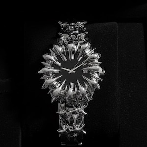 May include: A silver-toned wristwatch with a black face, presented in a black box. The watch features a unique, spiky design around the face and on the band, creating a bold, avant-garde aesthetic. The watch is a luxury item.
