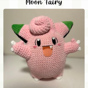 Könnte beinhalten: Ein rosa gehäkeltes Moon Fairy-Plüschtier mit grünen und schwarzen Akzenten an den Ohren, weißen Händen und Füßen sowie einer pfirsichfarbenen Zunge. Der Text "Crochet Pattern Moon Fairy" steht oben und "By Little Bunny Garden" unten.