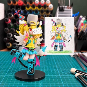 May include: A multi-colored anime-style figurine featuring a character with yellow hair, a gray top hat, white wings, and a blue tail, standing next to a matching hand-drawn illustration on paper that depicts the same character with a speech bubble.