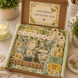 May include: A cardboard scrapbook surprise box filled with yellow and green floral paper ephemera, including die-cut butterflies, a bird, a camera, a teacup, and tags. The box features wooden letter tiles spelling HAPPINESS and two admit one tickets.