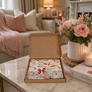 May include: An open cardboard box on a marble table, revealing a floral-patterned item wrapped in tissue paper and tied with a bow. The box is surrounded by a bouquet of pink roses, a candle, and a book.