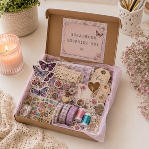 May include: A scrapbook surprise box filled with craft supplies. The box contains butterfly stickers, lace trim, decorative tape, and embellishments in shades of purple, pink, and cream. The box has a label that reads "Scrapbook Surprise Box."