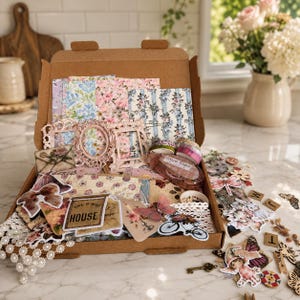 May include: A brown cardboard box filled with craft supplies. The box contains patterned paper, pink picture frames, decorative stickers, washi tape, and lace trim. The theme is vintage and floral, with a variety of embellishments.