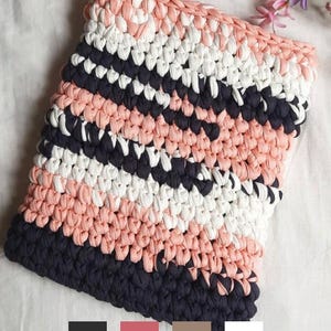 May include: A square, hand-crocheted pillow cover with a striped pattern. The stripes alternate between navy blue, peach, and white. The pillow cover is made of a thick, textured yarn, and the stitches are clearly visible.