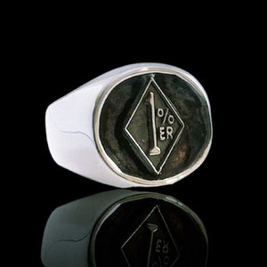Peut inclure: Une bague en argent avec une pierre ovale noire ornée d'un symbole blanc en forme de diamant avec le texte "1% ER" à l'intérieur.