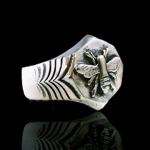 May include: A silver ring with a bee design. The ring has a textured band with a geometric shape.