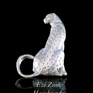 Puede incluir: Escultura de guepardo de plata, sentado con la cola enroscada alrededor de sus patas. La escultura está detallada con manchas y tiene un aspecto realista. La escultura está sobre un fondo negro con el texto "Ezi Zino Handmade Jewelry" debajo.