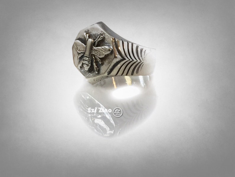 May include: A silver ring with a hexagonal design featuring a detailed bee sculpture. The band has a textured pattern.