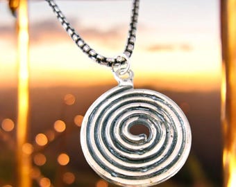 May include: A silver necklace with a round pendant featuring a spiral design. The pendant is hanging against a blurred background of golden lights.