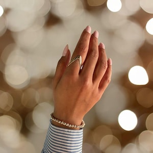 Puede incluir: Un anillo de plata con un diseño único y abstracto. El anillo se lleva en el dedo índice de una mano con esmalte de uñas rosa claro. La mano lleva una pulsera de cuentas plateadas.