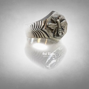 May include: A silver ring with a hexagonal design featuring a detailed bee motif. The ring has a textured band with a repeating pattern.