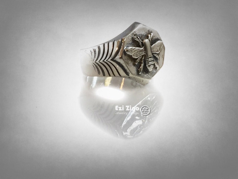 May include: A silver ring with a hexagonal design featuring a detailed bee motif. The ring has a textured band with a repeating pattern.