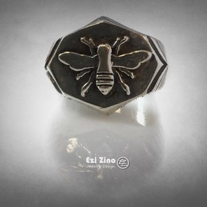May include: A silver ring with a hexagonal face featuring a raised bee design. The bee is detailed with wings, legs, and a segmented body.