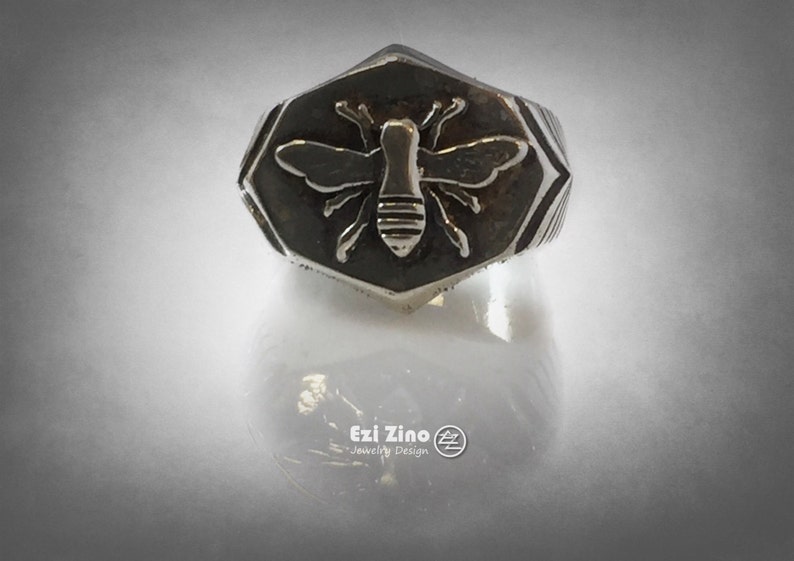 May include: A silver ring with a hexagonal face featuring a raised bee design. The bee is detailed with wings, legs, and a segmented body.