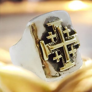Könnte beinhalten: Ein silberner Ring mit einem goldenen Jerusalem-Kreuz-Design. Der Ring ist quadratisch und hat eine strukturierte Oberfläche.